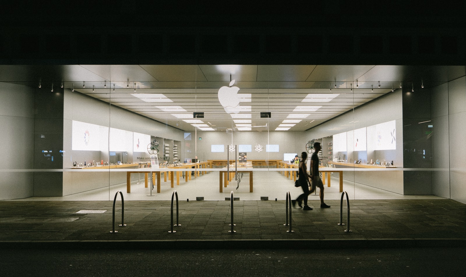An Apple Store storefront at night, with display tables visible through the glass and two people walking past