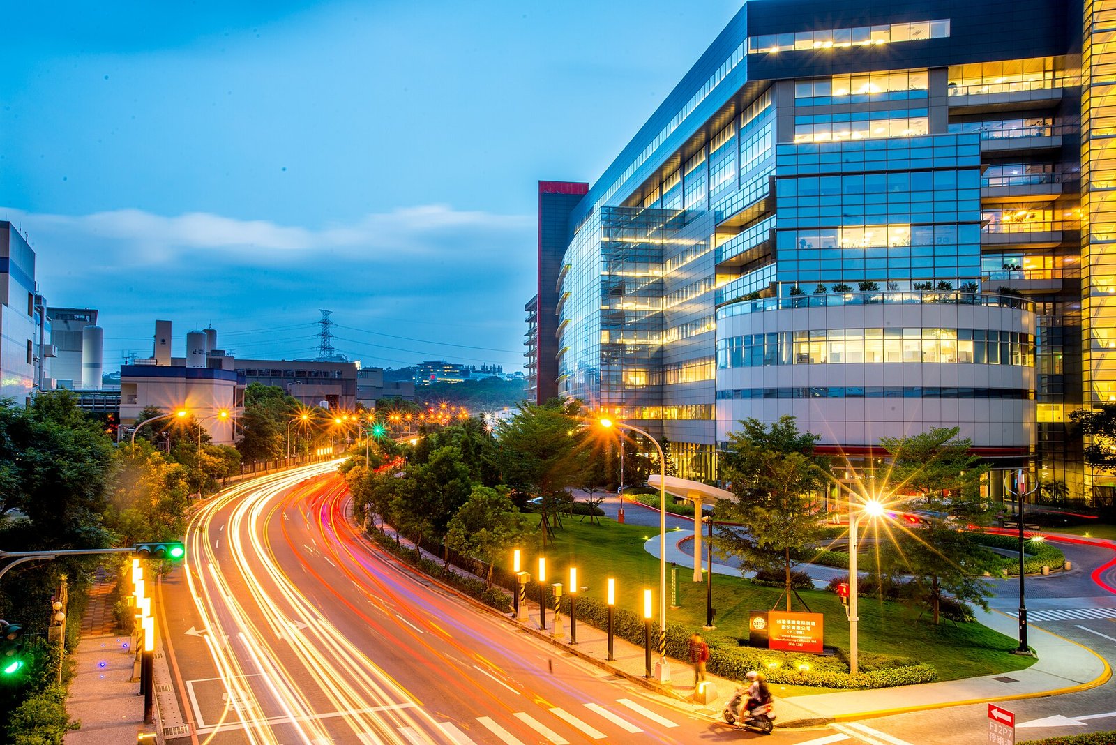 TSMC's Fab 12B building in the Hsinchu Science Park at dusk, with light trails from traffic on the road below, photographed in 2015; a contextual image of the foundry that manufactures the M6 and is expected to manufacture the A20 Pro.