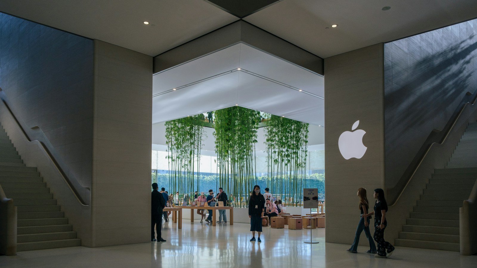 Interior of an Apple Store, with the Apple logo on the wall, wooden display tables, a bamboo grove behind glass and visitors between two stone staircases. Illustrative image.